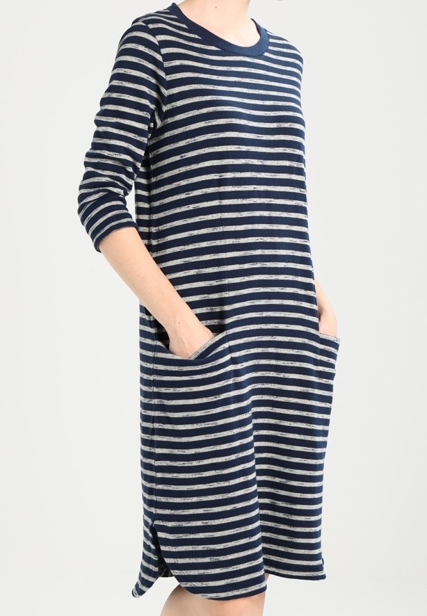 Femme portant une robe rayée horizontale bleu marine et blanc, longueur genou, manches longues et deux poches avant, mains glissées à l'intérieur.
