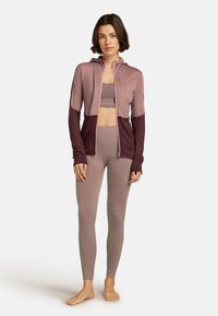 Donna in piedi a piedi nudi, con addosso leggings mauve, reggiseno sportivo abbinato e una felpa con zip bicolore mauve e bordeaux, con il cappuccio abbassato.