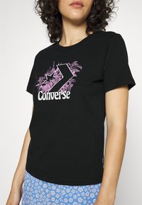 Svart bomulls-t-shirt med en stor rosa Converse-logotyp med en stiliserad pil och blommönster. Kort ärm med rund halsringning.