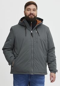Grå isolert jakke med hette, med brun fôr, glidelås foran og sidelommer. Glatt tekstur og justerbare snorer.