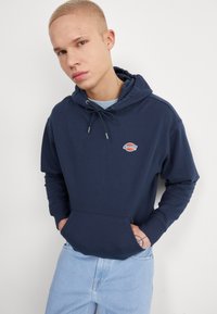 Felpa blu navy con tasca frontale, cappuccio regolabile con cordino e logo ricamato. Abbinata a jeans di denim azzurro. Texture liscia.