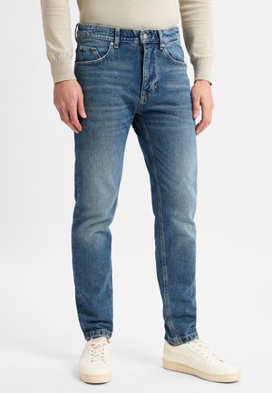 Mann in blauen Denim-Jeans, beigem Pullover und cremefarbenen Sneakers, der vor einem schlichten hellen Hintergrund steht.
