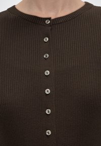 Κοντινό πλάνο καφέ ριμπ πλεκτού τοπ με επτά μικρά στρογγυλά κουμπιά κατά μήκος του κέντρου μπροστά και στρογγυλή λαιμόκοψη.