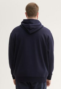 Marineblauwe hoodie met een zachte textuur, voorzien van een grote kap, geribbelde manchetten en een zoom. De achterkant toont een eenvoudig, onversierd ontwerp.