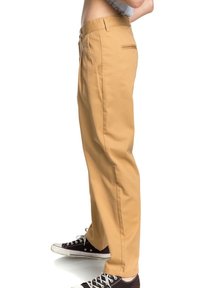 Khaki chinos s rovnými nohavicemi, vyrobené z tkané látky, s předními kapsami a zadní kapsou na zip. Spárovány s černými teniskami.