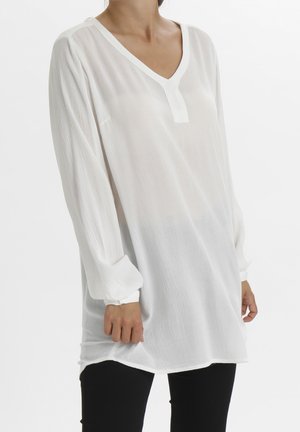 Tunique blanche à manches longues avec un col en V et un tissu texturé. Les manches sont froncées au niveau des poignets, créant une silhouette décontractée.