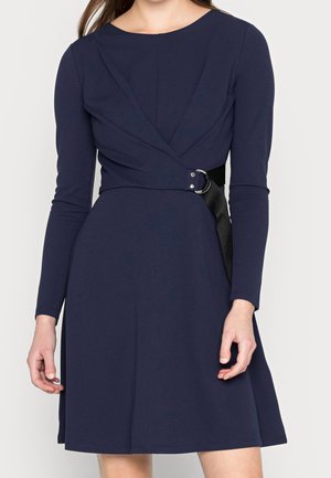 Robe longue bleu marine à manches longues avec un haut ajusté et une jupe évasée, présentant un design croisé et une ceinture noire avec une boucle argentée.