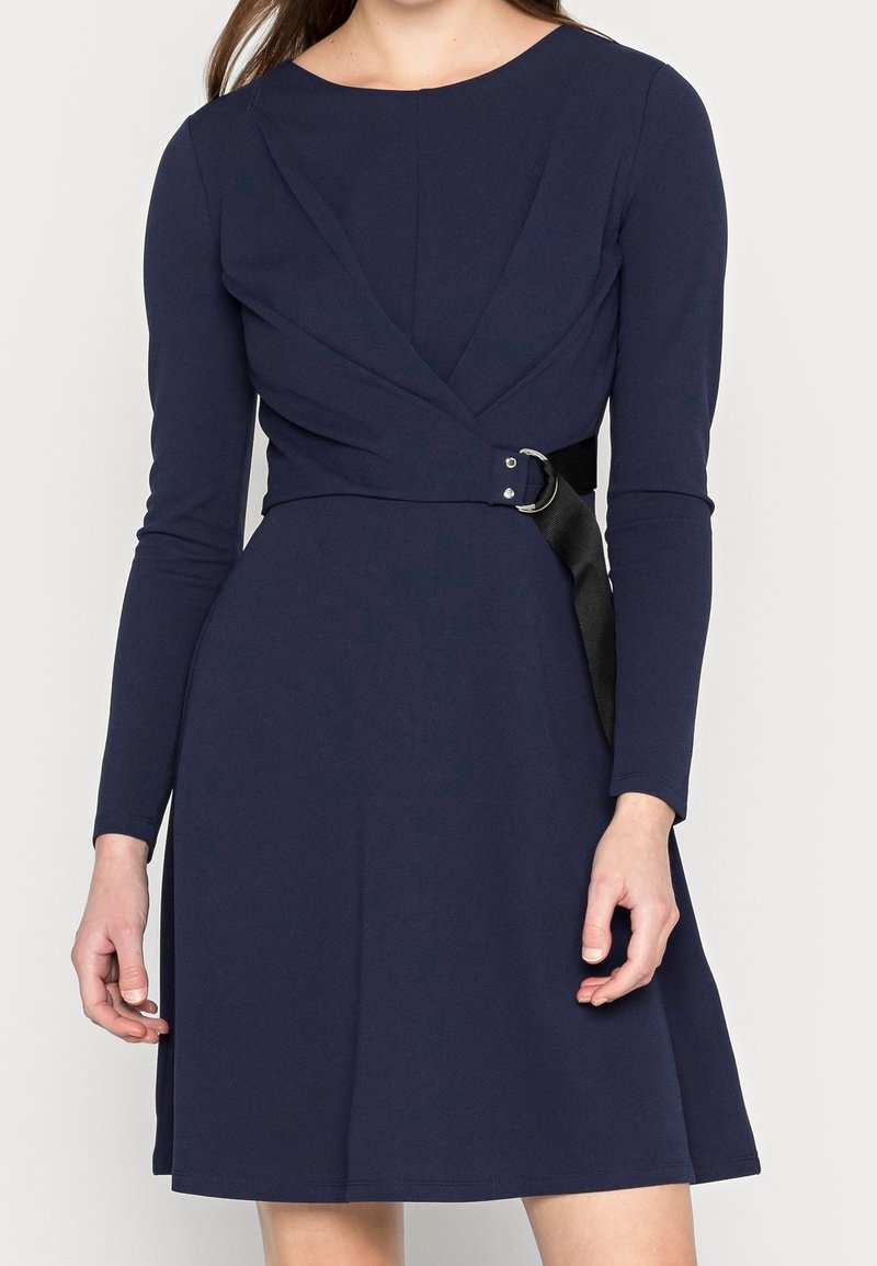 Mörkblå långärmad klänning med en figurnära bodice och en utsvängd kjol, med ett korsande mönster och ett svart bälte med en silvrig spänne.