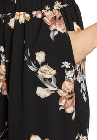 Tejido negro con flores grandes en tonos rosa y blanco, acompañado de hojas verdes. Incluye un bolsillo lateral y un detalle de cinturilla fruncida.