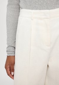 Cream-colored corduroy trousers with vertical ribbing, featuring a pleated front and a wide waistband. A hand is holding the waistband.