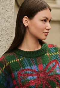 Zelený pletený sveter s festivalovým tartanovým vzorom a červenými, modrých a žltými akcentmi; veľký červený trblietavý detail mašle vpredu.