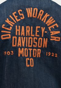 Ύφασμα από τζιν με πορτοκαλί κεντημένο κείμενο: "Dickies Workwear," "Harley-Davidson Motor Co," και τα έτη "1903" και "1922."