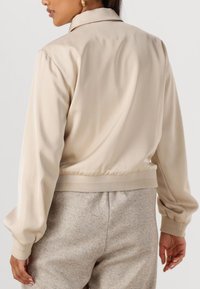 Beige bomberjack met een gekraagde halslijn en ribgebreide onderrand. Zachte, gladde textuur en opgezette mouwen. Draag met lichtgekleurde, gestructureerde broeken.