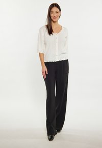 Cardigan bianco lavorato a maglia con maniche corte e chiusura a bottoni, abbinato a pantaloni neri a vita alta con gamba larga. Tessuto liscio e bordi scallop.