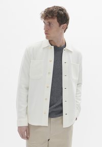 Lysfarvet skjortejakke med knaplukning, to brystlommer og en glat tekstur, lagd over en grå sweater. Enkel design, afslappet pasform.