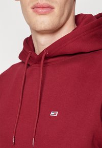 Sudadera de color marrón, hecha de una mezcla de algodón, con capucha ajustable, bolsillo tipo canguro y un pequeño logotipo en la esquina inferior izquierda. Textura suave.