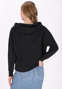 Felpa nera corta con scollatura ampia e grande cappuccio, realizzata in tessuto morbido. Abbinata a una gonna di jeans blu con tasche visibili.