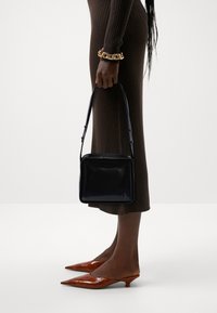 Borsa a tracolla in pelle verniciata nera con design piatto, dotata di manico superiore e logo in rilievo. Indossata con tacchi a punta marroni in pelle verniciata.