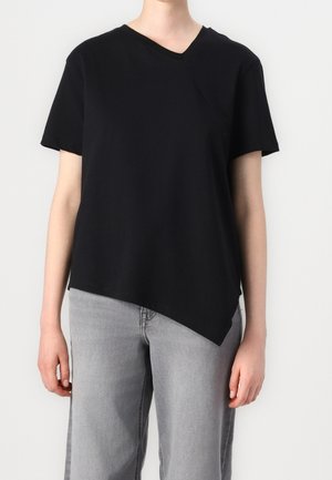 Persoon die een zwarte asymmetrische top met korte mouwen en V-hals draagt en lichtgrijze spijkerbroek tegen een effen lichte achtergrond.