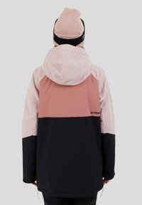 Persoon in een kleur geblokte jas met roze, stofroze en zwarte panelen en een roze beanie, wegkijkend tegen een effen grijze achtergrond.