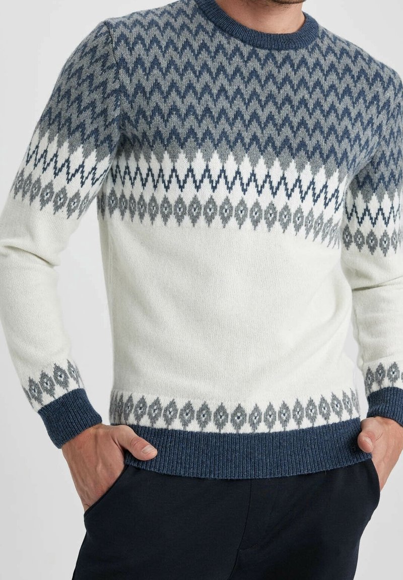 Homme portant un pull blanc avec des motifs géométriques en zigzag et diamant bleus et gris, mains dans les poches, sur fond clair uni.