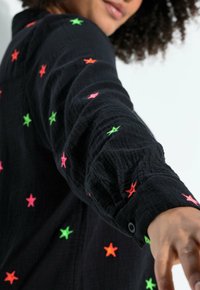 Persoon draagt een zwart shirt met lange mouwen met groene, rode en roze sterpatronen, arm naar voren gestrekt, gedeeltelijk gezicht en krullend haar zichtbaar.