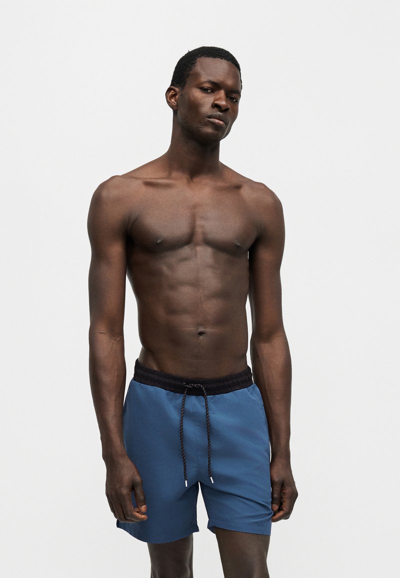 Άνδρας με σκούρο δέρμα χωρίς πουκάμισο, που φορά μπλε μαγιό με μαύρη μέση και κορδόνι, στέκεται μπροστά σε απλό φωτεινό φόντο.