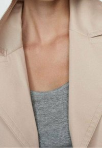 Trench-coat beige à large col, associé à un débardeur gris. Texture lisse, détails de couture minimaux sur le manteau.