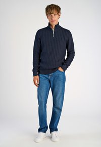 Marineblauer Kabelstrickpullover mit halbem Reißverschluss, kombiniert mit blauen Jeans und weißen Sneakers, mit entspannter Passform und strukturiertem Muster.