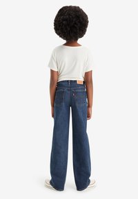 Džínové kalhoty s širokými nohavicemi v tmavě modré barvě s klasickým designem pěti kapes, viditelným šitím a zadní koženou nášivkou značky. Kombinované s bílou košilí.