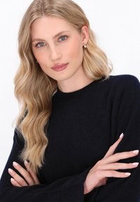 Blond model iført en marineblå sweater med rund hals, der viser en blød tekstur. Hun har let makeup og perlestuds som øreringe.
