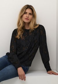 Blusa nera trasparente con texture floreale, maniche lunghe, chiusura con bottoni e dettagli a volants. Abbinata a jeans blu.