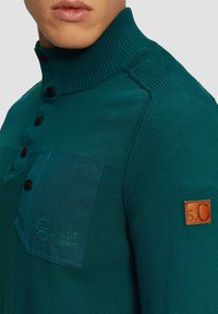 Türkis gestrickter Pullover mit hohem geripptem Kragen, ausgestattet mit einer kontrastierenden Brusttasche, schwarzen Knöpfen und einem Leder-Logo-Patch am Ärmel.