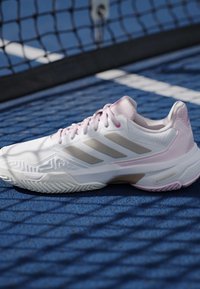 Hvid og lyserød Adidas tennissko på en blå baneoverflade med et net, der kaster skygger over skoen.