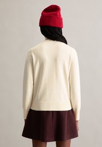 Eine cremefarbene Strickjacke mit langen Ärmeln, gerippten Bündchen und Saum, kombiniert mit einem dunkelburgunderfarbenen Rock. Eine rote Strickmütze wird getragen.