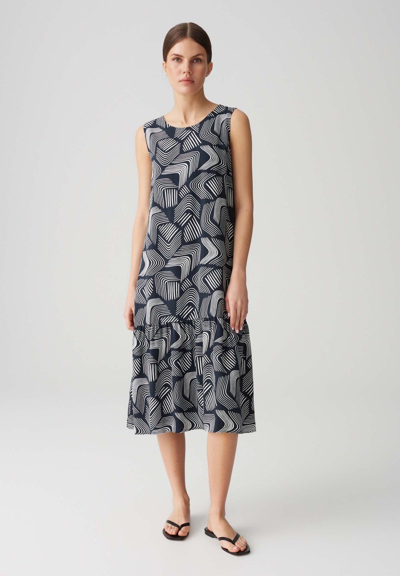 Marineblaues Kleid mit weißen abstrakten Wellenmustern. Ärmelloses Design, knielang, mit Rüschensaum. Getragen mit schwarzen flachen Sandalen.