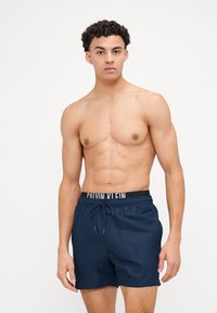 Pantaloni da nuoto navy con vita elastica e etichetta nera. Tessuto liscio con dettaglio del cordoncino; taglio sopra il ginocchio.