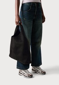 Person wearing wide-leg dark jeans and white sneakers holds a large black tote bag against a plain background.