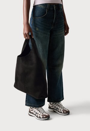 Een persoon in een wijde donkere spijkerbroek en witte sneakers houdt een grote zwarte tote bag tegen een effen achtergrond.