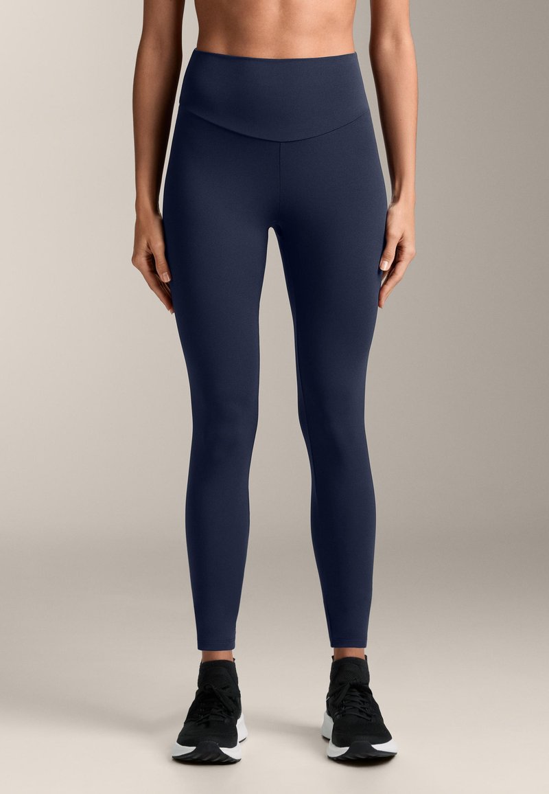 Leggings de culoare bleumarin, cu talie înaltă, realizate dintr-un material neted și elastic. Design ajustat cu cusături fără sărăcie și o bandă lată la talie, asortate cu pantofi sport negri.