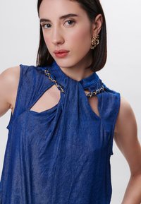 Top senza maniche blu con un collo annodato unico, caratterizzato da dettagli a taglio e accenti in catena dorata. Tessuto leggero e testurizzato.