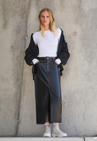 Zwarte neplederen maxi-jurk met een voorkleur, gecombineerd met een witte crop top en een oversized zwarte jas; witte hoge sneakers.