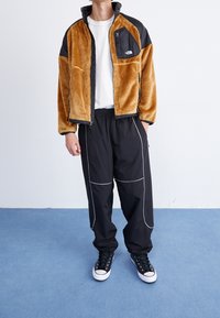 Giacca in pile marrone e nera con tasca con zip, abbinata a pantaloni neri con piping bianco e sneakers nere con suola bianca.