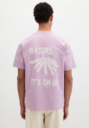 Mężczyzna w jasnofioletowej koszulce z białą grafiką stokrotki i napisem "Nature It's On Us" na plecach, stojący na jednolitym białym tle.