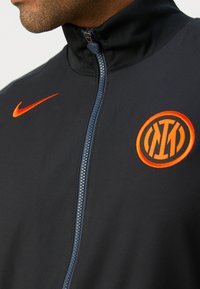 Crna sportska zip-up jakna od glatkog materijala, s istaknutim narančastim Nike logom i narančastim okruglim timskim grbom.