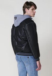 Giovane uomo che indossa una giacca bomber nera con cappuccio grigio e jeans azzurri, in piedi con un leggero angolo di torsione verso il lato contro uno sfondo neutro.
