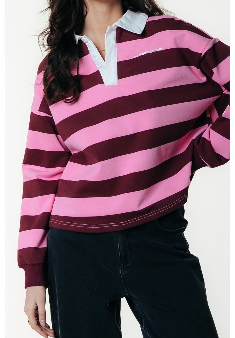 Femme portant un polo à manches longues avec de larges rayures horizontales roses et rouge foncé et un pantalon foncé sur un fond uni.