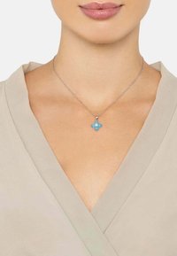 Srebrny naszyjnik z kwiatowym wisiorkiem ozdobionym czterema niebieskimi kamieniami szlachetnymi. Łańcuszek jest delikatny, a wisiorek znajduje się na środku dekoltu.