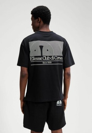 Mężczyzna ubrany w czarną koszulkę i spodenki Ellesse Club di Corsa, widziany od tyłu na jednolitym jasnoszarym tle.