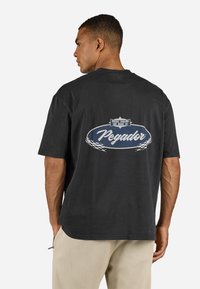 Sort bomulds t-shirt med et stort grafisk tryk på ryggen, der har teksten "Pegador" og et ternet mønster, afslappet pasform, korte ærmer.